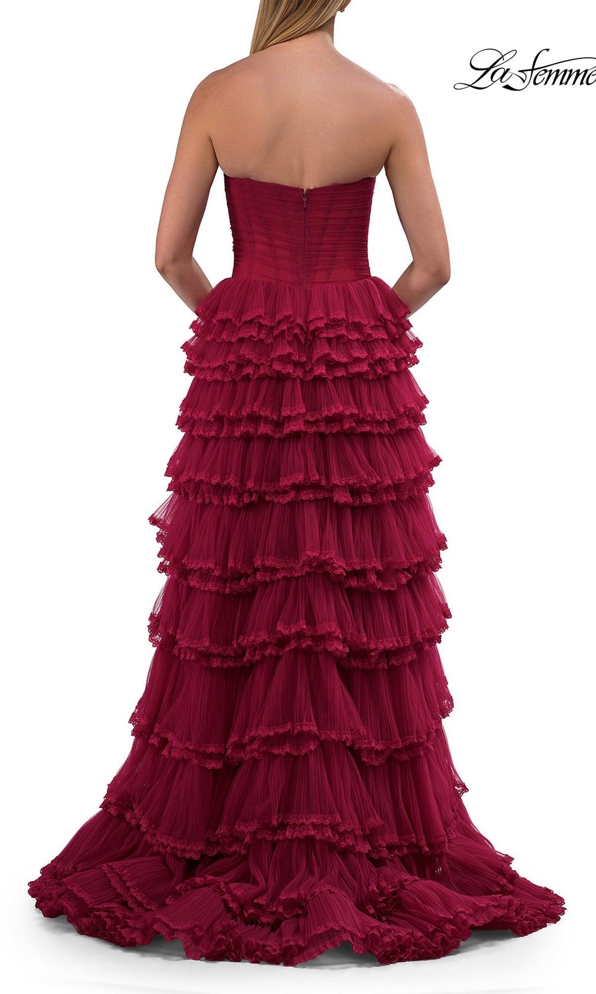 A woman stands facing away from the camera, wearing the La Femme 33727 long prom dress—a strapless, floor-length burgundy gown featuring a corset bodice and multiple layered ruffles.